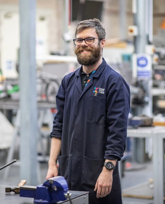 A lecturer in a navy-blue Glasgow Kelvin College coat standing confidently in a mechanical engineering workshop, surrounded by workbenches and tools. A lecturer in a navy-blue Glasgow Kelvin College coat standing confidently in a mechanical engineering workshop, surrounded by workbenches and tools.